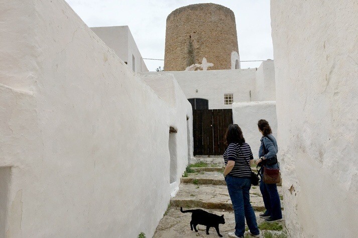 Tourists exploring cobbled alley in Ibiza with windmill and black cat